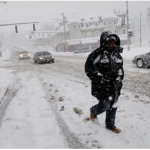 A person bundled up in warm clothing, walking briskly down a snowy city street in Syracuse, with snow-covered trees and buildings in the background.